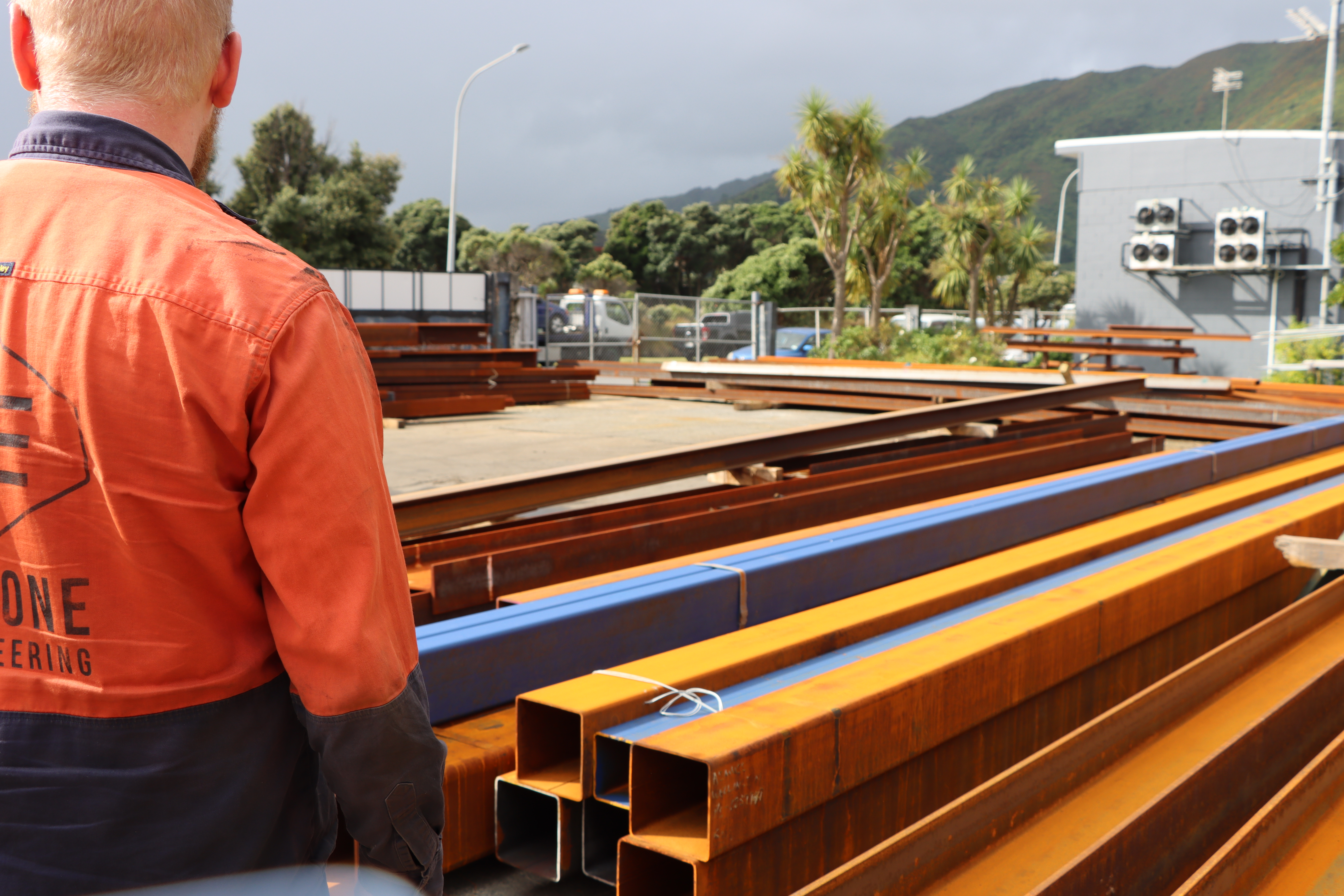 Kieran Houssenloge, a pakeha male wearing a high-vis vest, is standing next to stacked raw steel for the build in the yard.