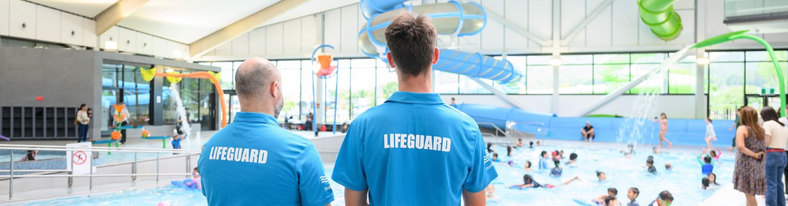 Two lifeguards stand with their backs to the camera watching over children playing in a pool banner image