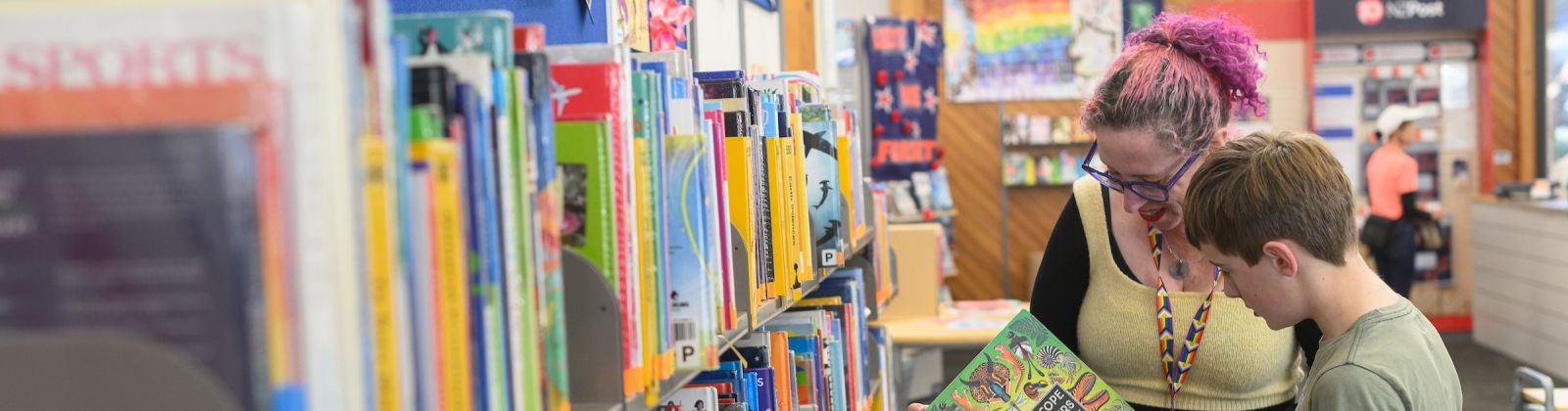 A librarian and child look at book while standing next to a row of library shelves banner image