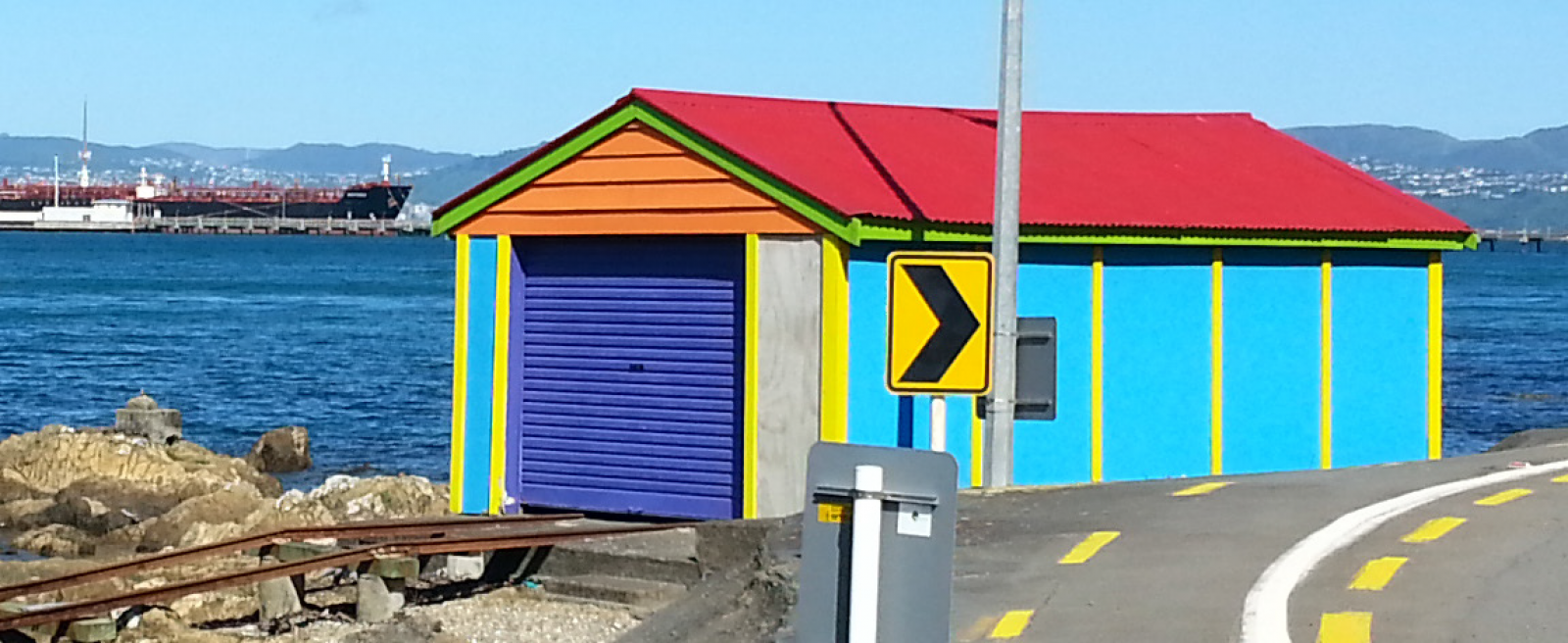 A brightly painted boat shed between the road and beach in Lowry Bay banner image